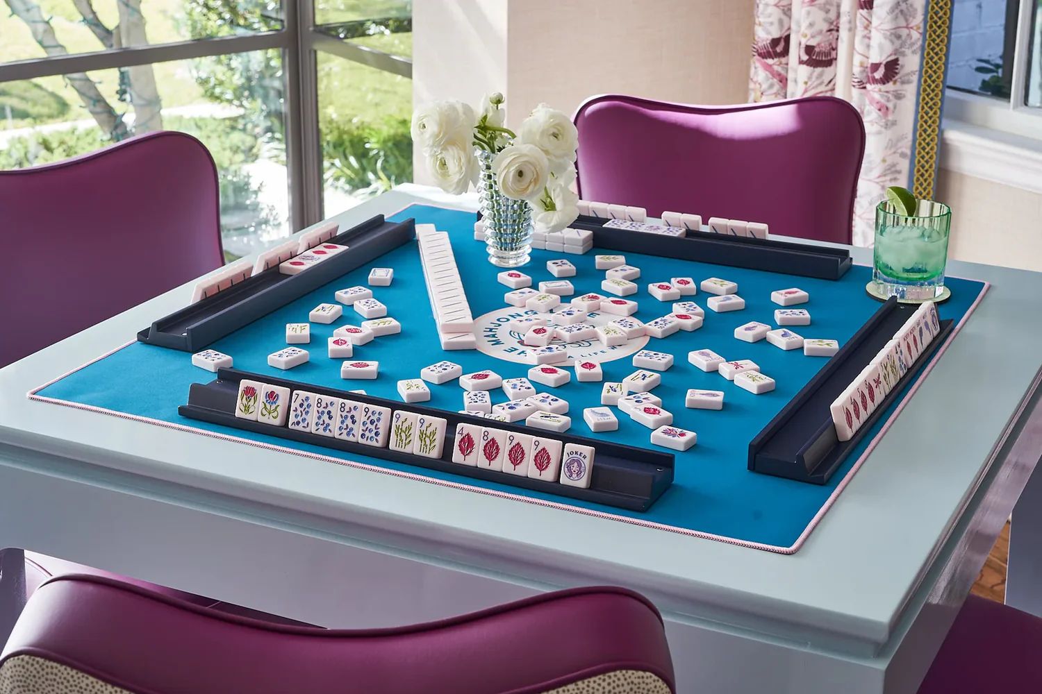A mahjong table set on blue felt with fresh flowers and a cocktail